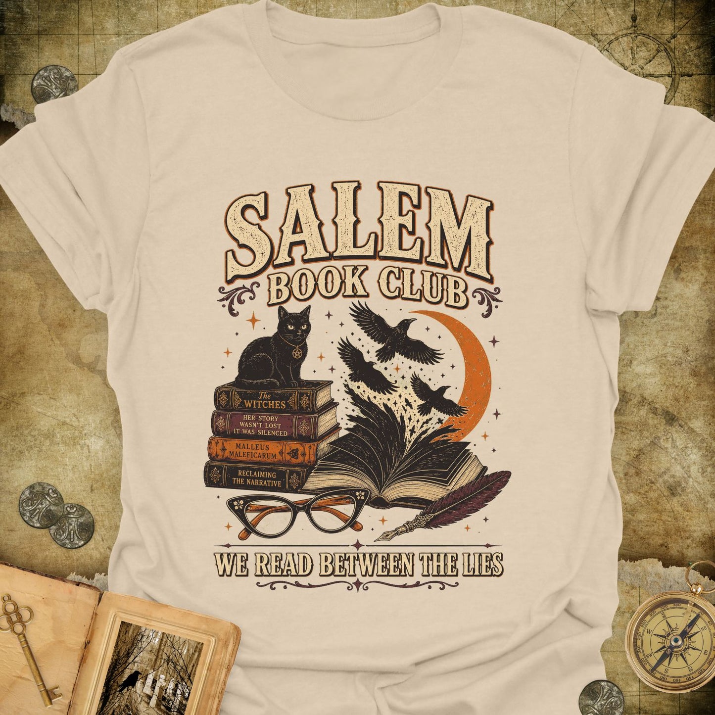 A cream-colored graphic t-shirt featuring a 'Salem Book Club' design with a black cat, open books, crows, glasses, and a compass, displayed on a textured vintage map background.