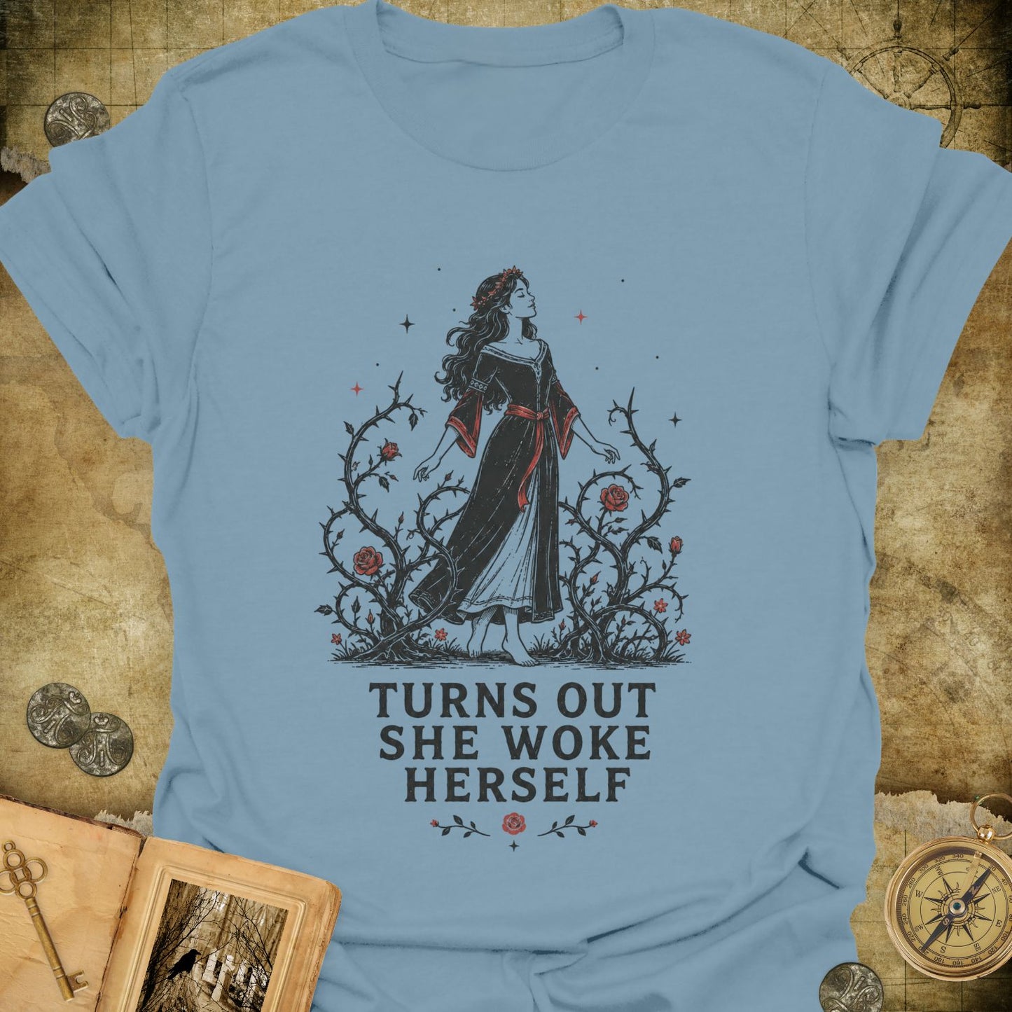 A light blue graphic t-shirt laid flat on a textured, vintage-style parchment background. The graphic features an illustration of a woman in a black and red dress surrounded by thorny branches, with the text 'TURNS OUT SHE WOKE HERSELF' printed below in a bold serif font.
